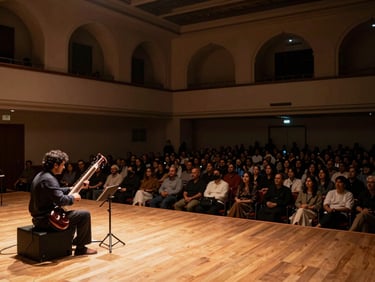 Panoramic wide shot of an intimate mehfil concert hall, a sitarist seated on a low stage, warm amber footlights, sparse attentive audience in shadow, no phones visible, architectural arches in the background