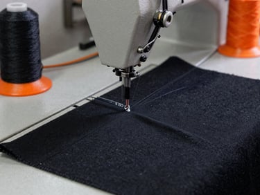 Close-up of heavy-duty black textile being stitched on an industrial sewing machine, strong workshop lighting, focus on durable thread, industrial charcoal and safety orange tones, 35mm lens.