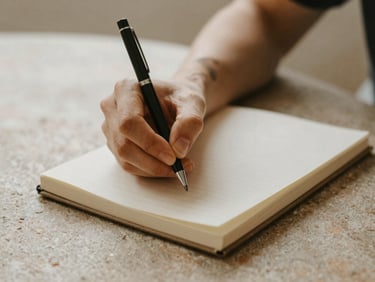 Close-up of hands writing in a linen journal on a warm stone table, soft morning light, shallow depth of field.