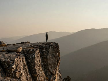A solitary figure standing on a vast stone cliff overlooking a misty mountain valley at quiet dawn, muted earthy tones, 35mm film texture.