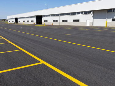 A newly completed asphalt parking lot and loading bay of a modern industrial warehouse, with a yellow road marking machine painting lines