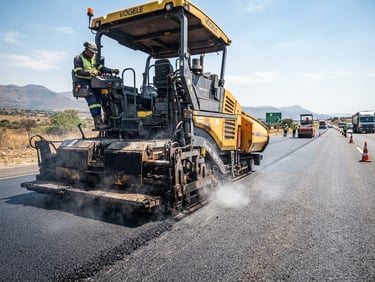 Low-angle heroic shot of a heavy paving machine laying hot-mix asphalt on a highway under bright South African daylight, rich charcoal and safety yellow tones