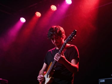 Low-angle shot of a guitarist on a dark stage under saturated red and pink spotlight, sweat and dust visible in the air, cinematic grain.