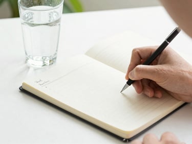 A person writing down a meal plan in a clean notebook next to a glass of water, bright daylight