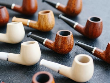 Close-up editorial photograph of a selection of finely crafted wooden smoking pipes, showcasing grain and finish, on a dark, textured surface.