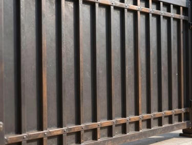 Close-up architectural shot of a custom-fabricated bronze-accented metal fence and gate for a private estate, clean welded seams, high-contrast sunlight casting linear shadows, industrial premium finish
