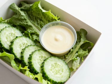 Overhead view of a fresh garden salad box, crisp mixed greens, sliced cucumbers, and a small cup of house dressing.