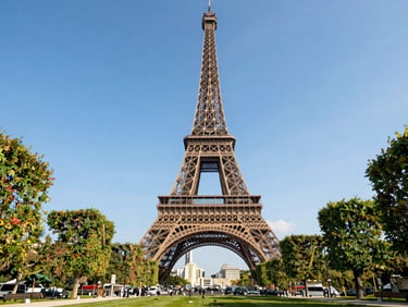 Eye-level clean shot of the Eiffel Tower framed by manicured trees under crisp afternoon sun, high-end travel editorial style.