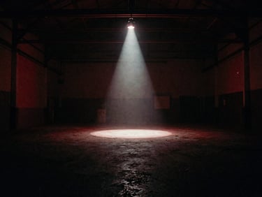 Wide atmospheric shot of a dimly lit empty warehouse venue with a single harsh spotlight cutting through haze, low-key ambient red glow, grainy 35mm film