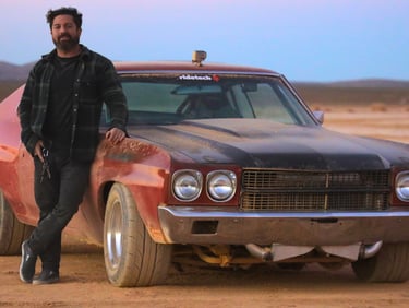 A man leans against a classic red and black muscle car in the desert at sunset.
