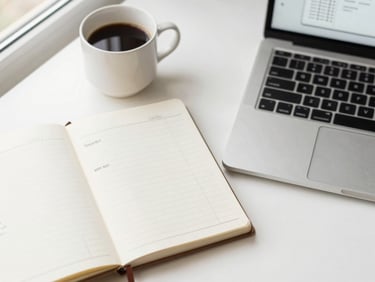 Overhead flat-lay of a notebook with travel prompts, a coffee cup, and a laptop showing itinerary drafts, soft warm tones, clean modern workspace.