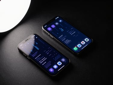 Overhead flat-lay of two smartphones displaying dark mobile app interfaces with blue and violet data visualizations, cold white studio light, matte black surface, no text visible, high contrast product photography