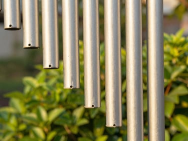 Close-up of hand-brushed aluminum chime tubes catching warm golden hour sunlight, lush green garden blurred in the background, shallow depth of field, 50mm lens.
