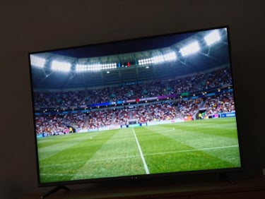 Angled close-up of a modern smart TV displaying a live sports broadcast, vibrant stadium lights illuminating a dark room, cinematic ambient glow.