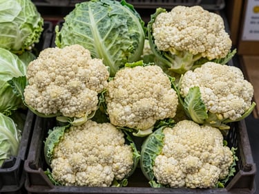 Crisp heads of cauliflower and green cabbage stacked in a wholesale market crate, natural overcast daylight, focus on texture.