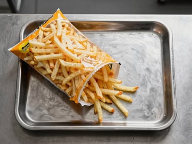 Overhead straight-on shot of a commercial kitchen prep table, a large bag of frozen french fries open beside a stainless steel tray, cool studio lighting, clean neutral background, no styling