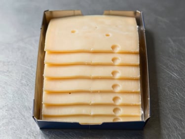Straight-on flat lay of individually wrapped cheese slices stacked in an open commercial carton on a clean metal surface, cool neutral studio lighting, no food styling