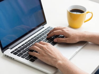 Hands typing on a clean modern laptop next to a cup of coffee, bright morning daylight