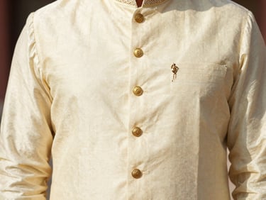A close-up of a premium cream-colored raw silk sherwani with gold button details and fine collar embroidery, warm daylight