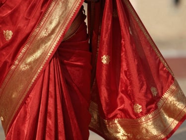 Detailed close-up of rich crimson silk saree fabric with gold zari embroidery patterns, warm side lighting, shallow depth of field