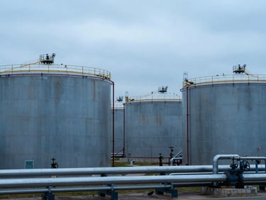 Wide-angle industrial petroleum storage facility, cylindrical steel tanks receding in cool blue-grey light, pipeline infrastructure in foreground, overcast sky above, no people, documentary framing