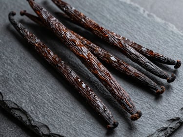 Overhead close-up study of whole vanilla beans arranged on a dark slate surface, natural daylight, rich brown tones, visible texture and oil sheen on pods, no people, no labels