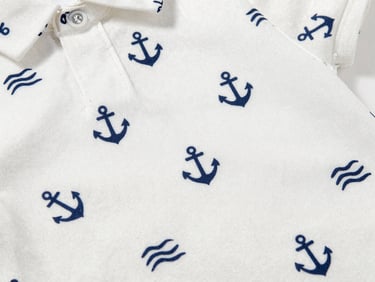 Close-up flat-lay of a children's shirt fabric with a small repeating maritime print — anchors and waves in navy on white jersey — showing texture and print sharpness, white background, studio light