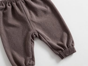 Flat-lay overhead of a pair of baby harem pants in French terry fabric on white surface, close-up of gathered ankle seam and waistband elastic, neutral studio light