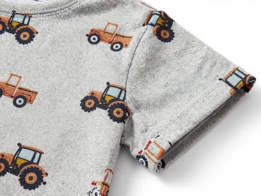 Close-up flat-lay of a children's shirt fabric with a small tractor and truck print in earthy tones on light grey jersey, showing print detail and weave texture, white background, diffuse studio light