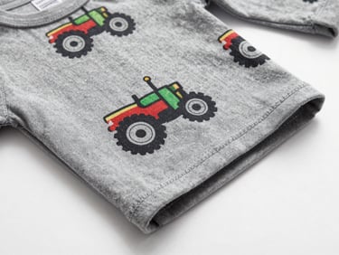 Flat-lay overhead of a folded long-sleeve toddler shirt in jersey fabric on a pure white surface, close-up showing a tractor print motif, soft studio light, seam detail visible