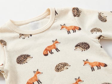 Close-up flat-lay of a baby shirt fabric showing a small animal print — foxes and hedgehogs in muted warm tones on off-white jersey — texture and print clarity visible, white background, soft studio light