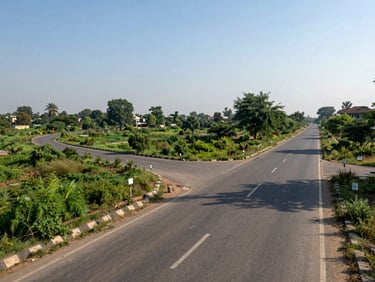 Wide-angle view of residential land near Delhi Road Hisar, showing connecting asphalt road, green trees in the background, and clear sunny sky.