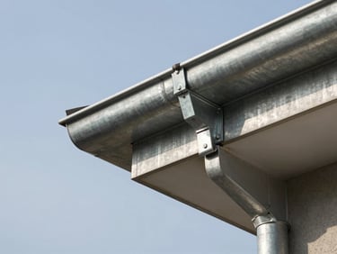 Close-up of a galvanised steel gutter being secured to a fascia board with brackets, new downpipe visible at the right edge, clear sky