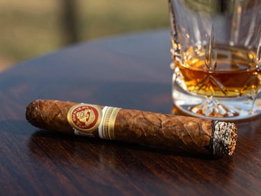 Close-up of a premium cigar with a gold embossed band resting on a dark walnut table next to a crystal rum tumbler