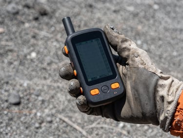 A rugged hand wearing a soiled protective glove holding a technical telemetry GPS terminal displaying coordinate grids, harsh midday sun, ash-covered ground in the background, tight environmental framing.