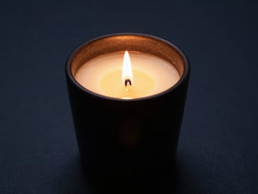 A hand-poured scented candle in a dark ceramic vessel, flame glowing softly, side-lit, warm golden wax, 35mm