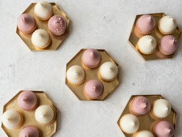 Elegant overhead flat lay of dusty rose and cream wedding sweets arranged in geometric gold trays on a textured plaster backdrop, soft morning light