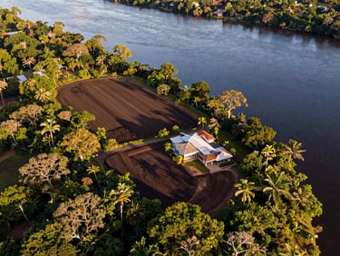 Aerial view of a riverfront estate in Moju, Pará, showing rich dark soil, native forest borders, and calm river waters under afternoon sun.
