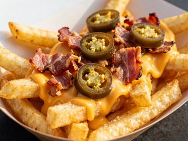 Close-up of golden crispy loaded fries topped with melted cheese sauce, chopped bacon, and sliced jalapeños in a paper food truck tray, vibrant editorial food photography