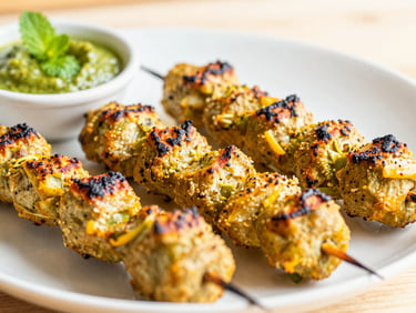 Close-up of freshly grilled seekh kababs on a platter with mint chutney, bright natural kitchen light, vibrant saturated colors, shallow depth of field.