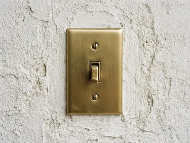 Bright architectural photography, straight-on macro shot of a premium brushed brass light switch mounted on textured off-white plaster, soft natural daylight, 35mm