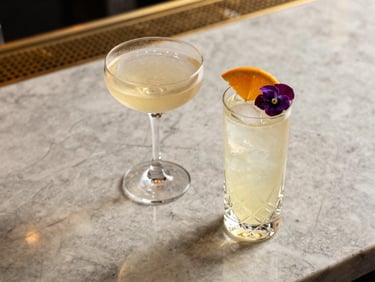 Overhead view of two signature cocktails on a polished marble bar top, champagne coupe and a crystal highball, fresh citrus garnish and edible flower, golden-hour ambient light