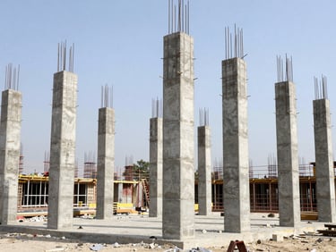 Wide environmental shot of an active Indian construction site, concrete pillars rising under bright daylight, high-contrast industrial tones.