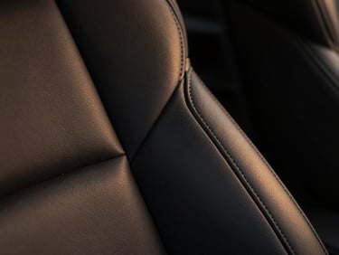 Close-up macro shot of premium black leather stitching on a luxury car seat, soft warm amber side lighting, shallow depth of field, high-end texture.