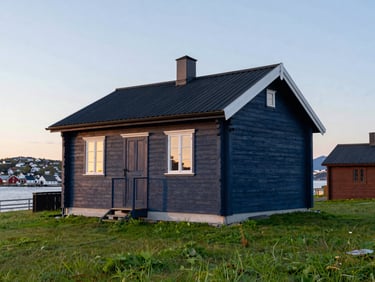 A well-maintained wooden Norwegian cabin in Vadsø under a soft golden midnight sun sky, clean green grass in the foreground, crisp details