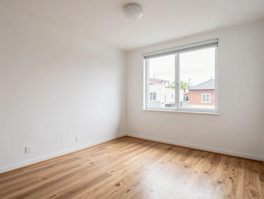 Living room of a compact two-bedroom apartment — wide angle showing layout and light, white walls, laminate flooring, window to street view