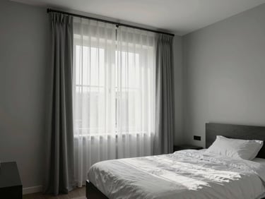 Bedroom interior with double bed, large window with sheer curtain letting in morning light, light grey walls, minimal decor