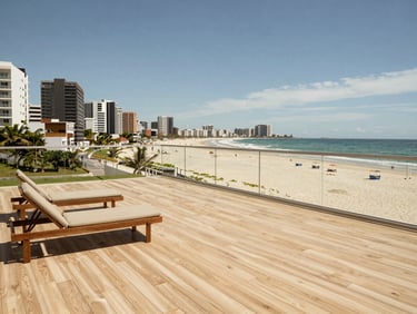 Wide-angle shot of a beachfront apartment terrace in daylight, light wood decking, two loungers, ocean horizon visible beyond a glass railing, Brazilian coastal town