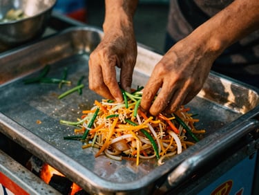 Intimate close-up of hands preparing fresh pad thai on a metal cart, glowing embers underneath, natural night lighting