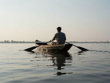 A boatman on a wide haor channel at midday, paddle resting across the bow, the flat water mirroring a pale sky, shot from low and slightly behind so the figure is silhouetted against the water's surface